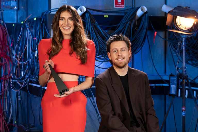 Comedians Melanie Bracewell in a red outfit holding an ARIA Award and Tim McDonald in a brown suit posing backstage amid studio cables